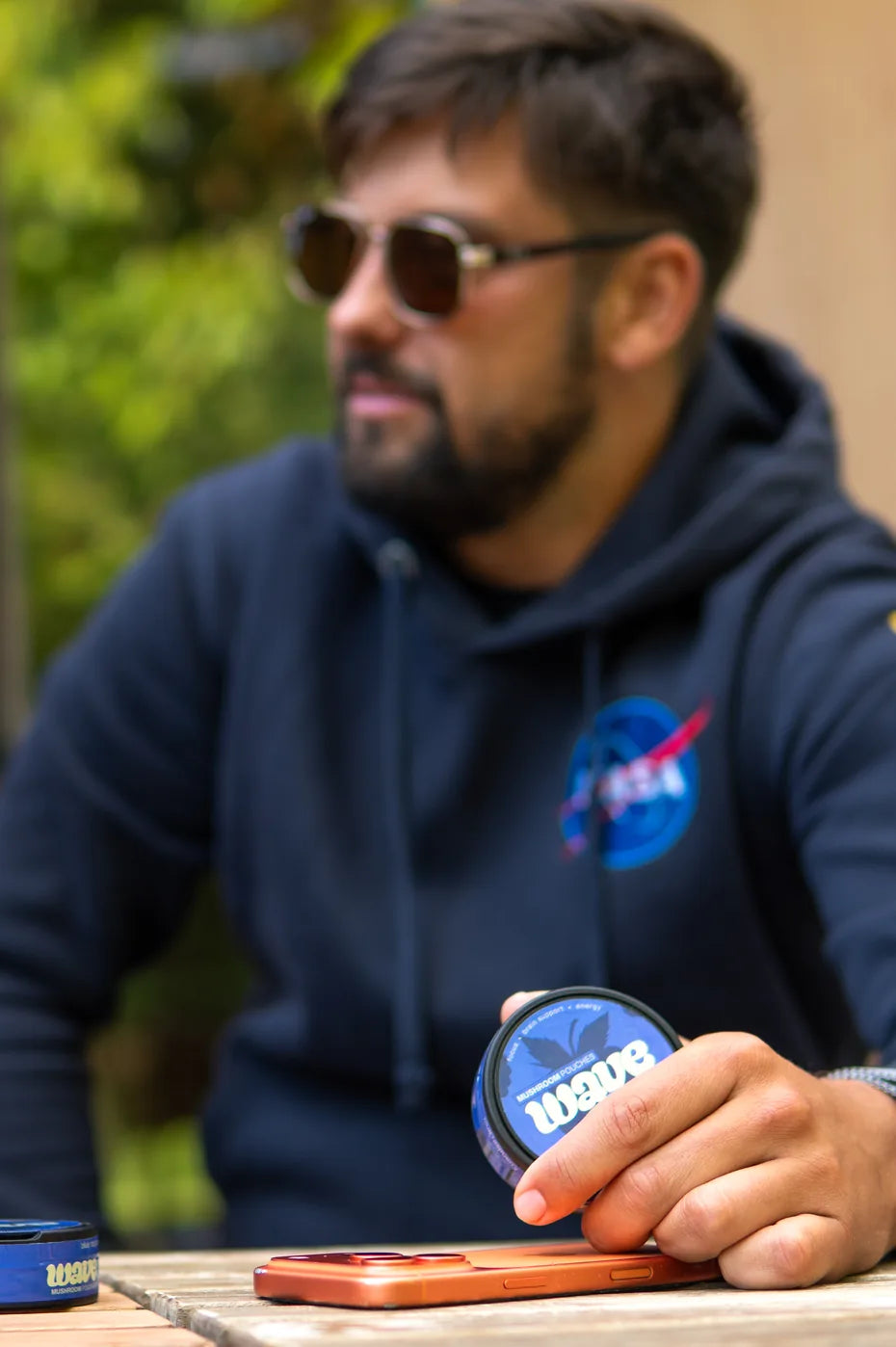 Man met zonnebril en marineblauwe hoodie aan een tafel buiten, met een blikje WAVE in zijn hand